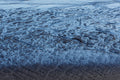 Shallow seawater receding across black volcanic sand, creating intricate blue, silver and dark rippled patterns along the shoreline.