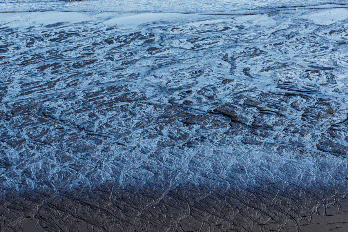 Shallow seawater receding across black volcanic sand, creating intricate blue, silver and dark rippled patterns along the shoreline.