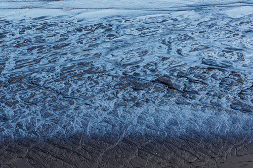 Shallow seawater receding across black volcanic sand, creating intricate blue, silver and dark rippled patterns along the shoreline.
