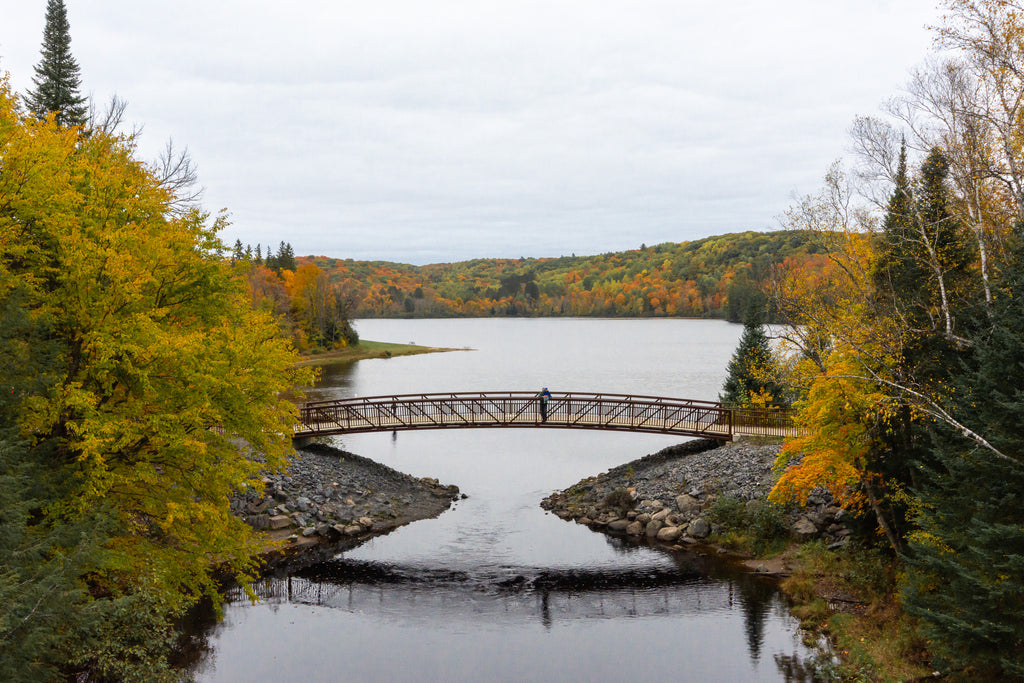 An adult holding a small child pauses at the centre of a bridge spanning a calm lake, surrounded by the autumn colours of Algonquin Park.