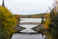 An adult holding a small child pauses at the centre of a bridge spanning a calm lake, surrounded by the autumn colours of Algonquin Park.