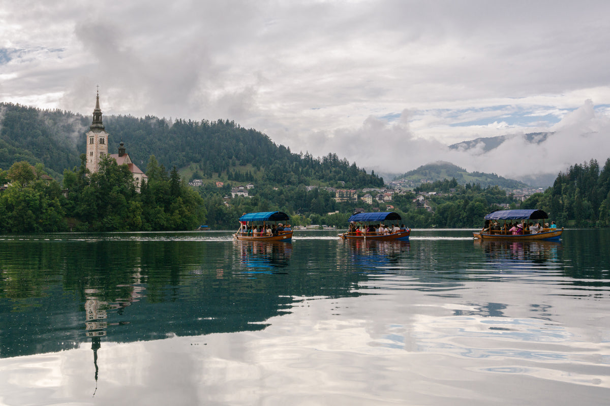 Three covered boats cross a reflective lake beneath a cloudy sky, with a church and forested hills in the distance.