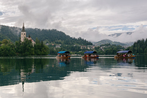 Three covered boats cross a reflective lake beneath a cloudy sky, with a church and forested hills in the distance.