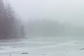 A frozen lake covered in dense fog, with bare trees fading into the mist along the left shoreline and scattered branches resting on the snow-covered ice.