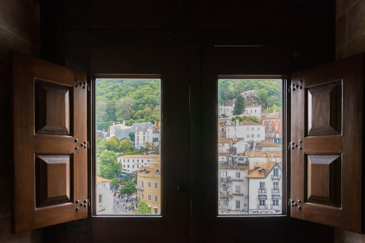 Open wooden shutters frame two tall windows overlooking the tiled rooftops and green hillside of Sintra from inside the National Palace.