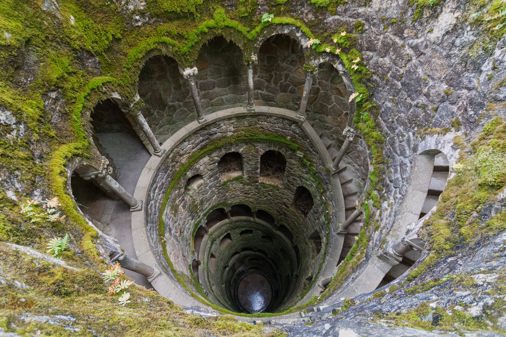 View looking down into the Initiation Well at Quinta da Regaleira, where a moss-covered spiral staircase and repeated stone arches descend into darkness.