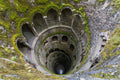 View looking down into the Initiation Well at Quinta da Regaleira, where a moss-covered spiral staircase and repeated stone arches descend into darkness.
